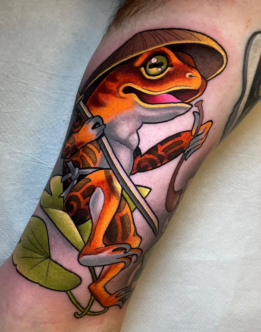 Tatuaje Neotradicional de una rana fumando pipa con un sombrero vietnamita y llena de tatuajes. Está sentada en una hoja de ginko mientras sujeta un sable. Tatuaje realizado a color (verdes, naranjas y amarillos) por Helena Ayala