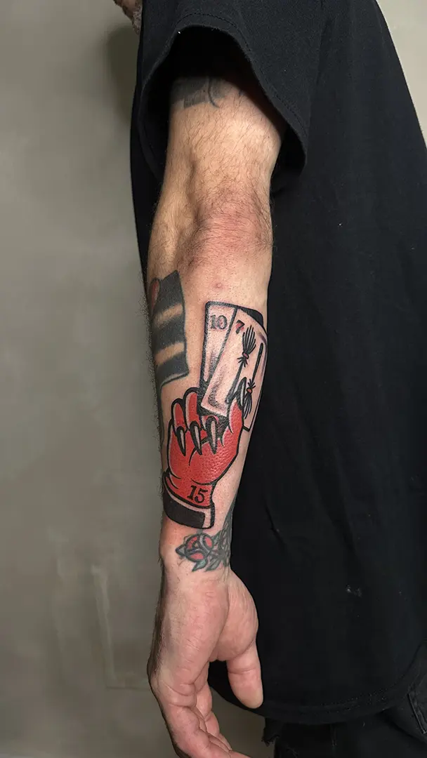 Tatuaje estilo Tradicional de la mano de un diablo (roja) con dos cartas en la mano (el 10 y el 7). El diablo tiene tatuado el 15 en la muñeca. Realizado en Karratu Tattoo
