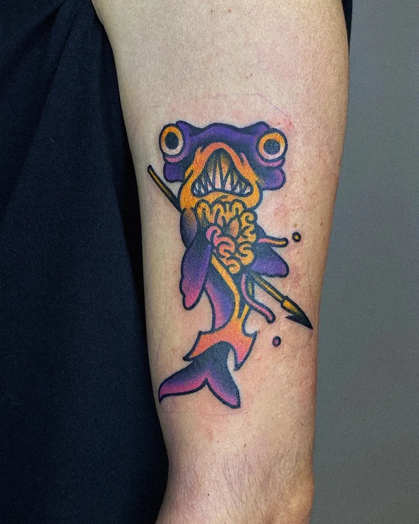 Tatuaje de tiburón hecho en estilo tradicional con un arpón en tonos morado, rosa y amarillo, realizado por Don Ander De Órganos en Karratu Tattoo Estudio de Tatuajes.