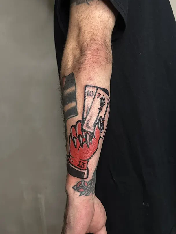 Tatuaje estilo Tradicional de la mano de un diablo (roja) con dos cartas en la mano (el 10 y el 7). El diablo tiene tatuado el 15 en la muñeca. Realizado en Karratu Tattoo